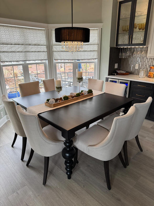 Dining room with a black table and white chairs, large windows, and kitchen in the background.